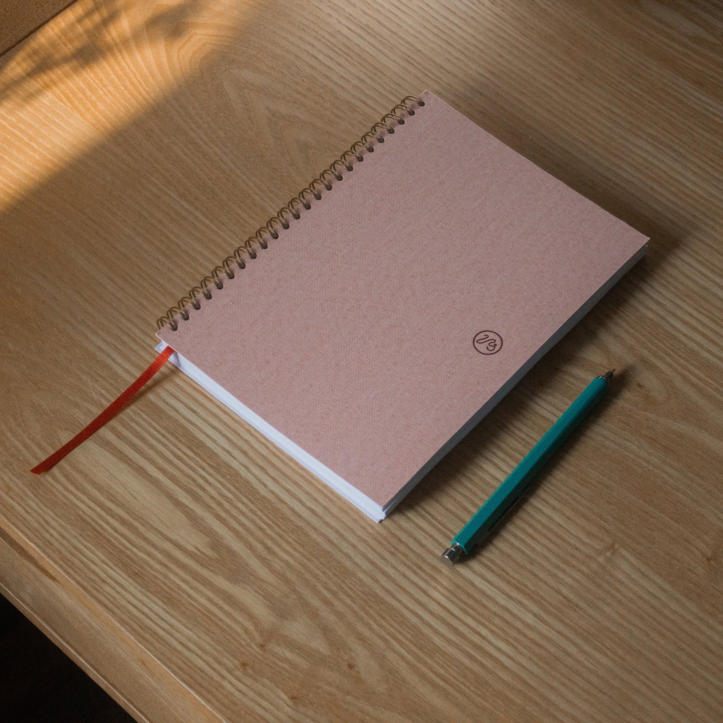 Pink spiral-bound notebook with a green pencil on a wooden surface