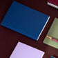 Blue notebook on a dark surface with other notebooks and a pen.